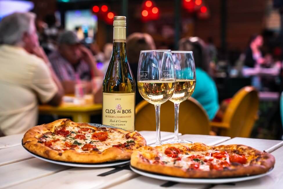 A bottle of Clos du Bois Chardonnay with two glasses of wine and two pizzas placed on a table in a casual, lively setting.