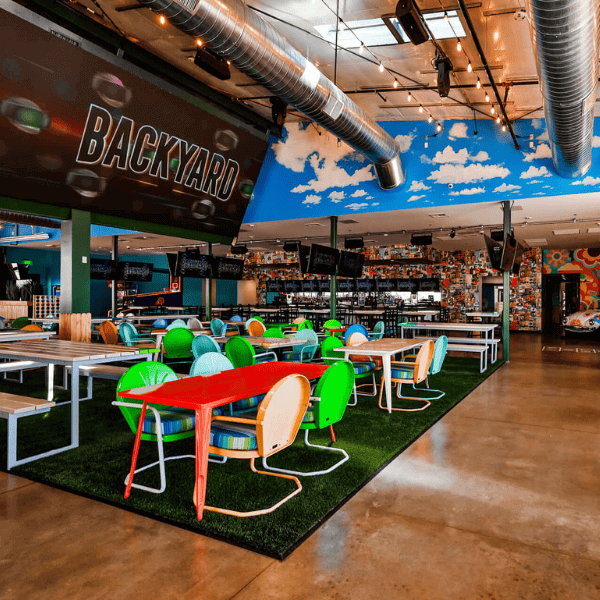 Vibrant indoor dining space with colorful retro chairs, turf flooring, and a large “Backyard” sign under a painted sky ceiling.