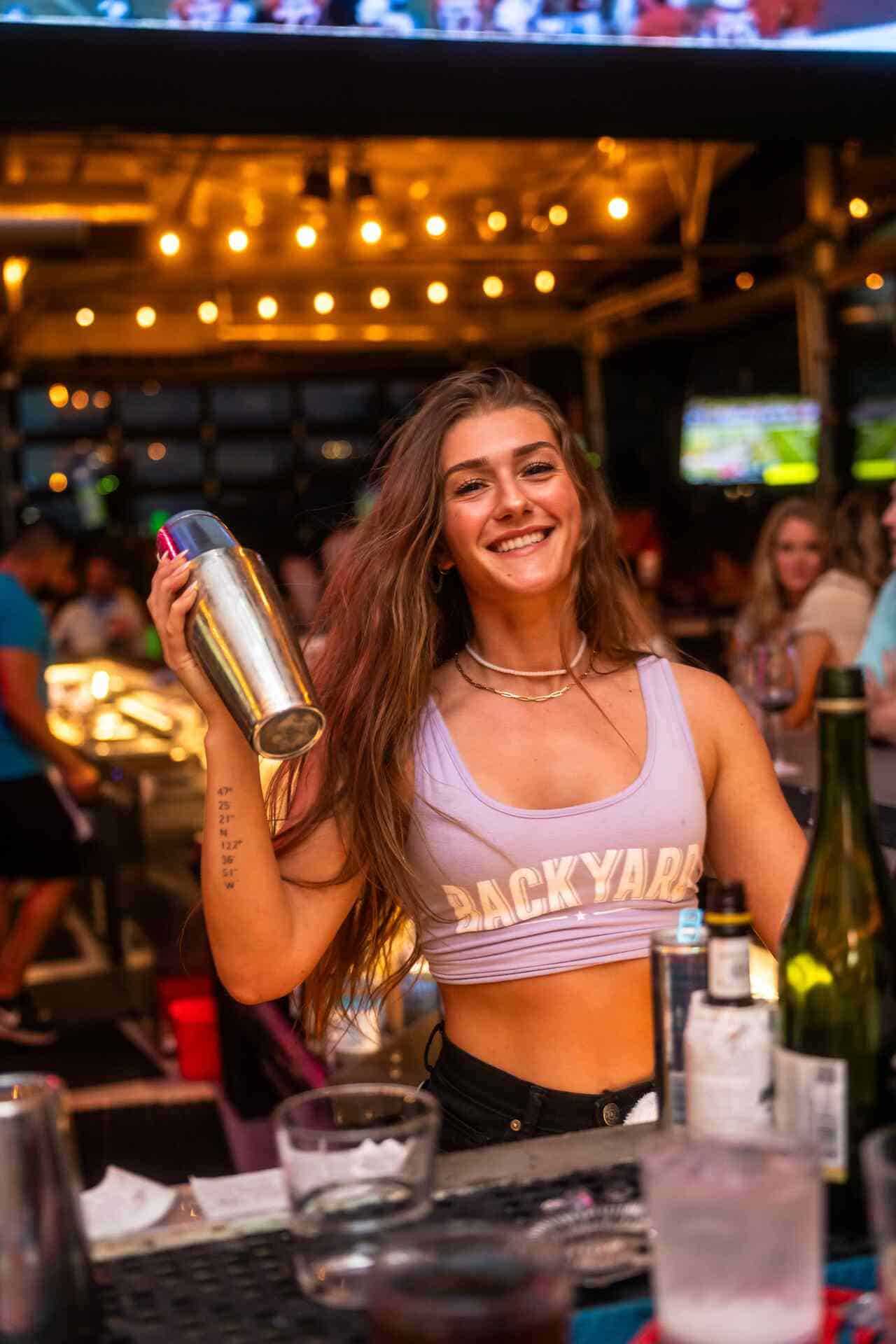 A bartender with long blonde hair pours a pink cocktail into a glass over ice at a bar with bright lights and TV screens in the background.
