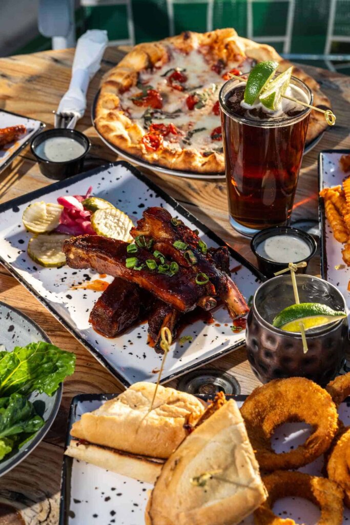 Flat-lay of a vibrant outdoor dining table with barbecue ribs, pizza, onion rings, cocktails, and a sandwich platter.
