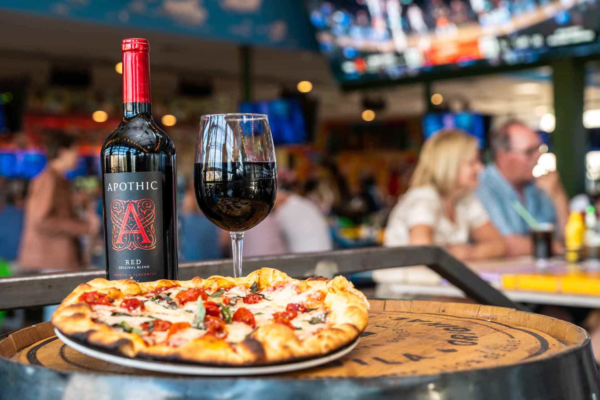 Glass and bottle of red wine with a freshly baked pizza on a wooden barrel table, guests dining in the background.