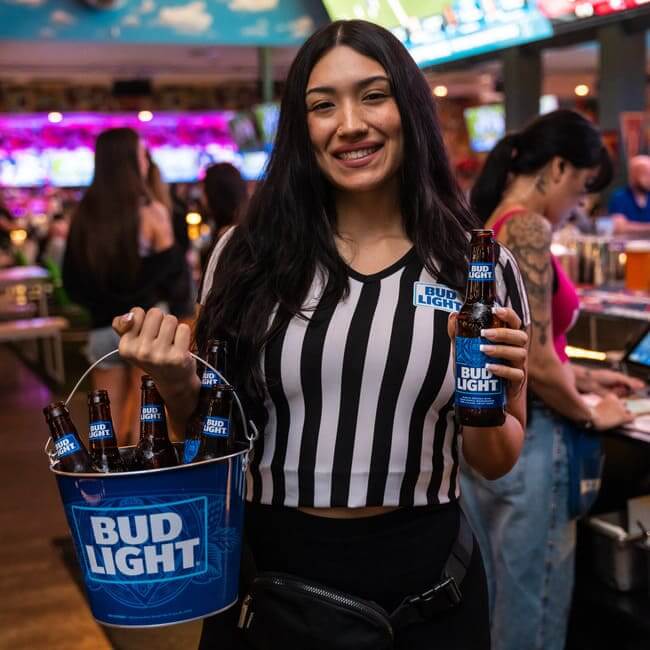 Server holding a bucket of Bud Light bottles and a beer inside a busy sports bar.