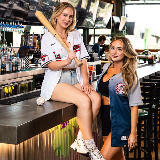 Two women in baseball jerseys posing in a bar, one holding a baseball bat and sitting on the counter