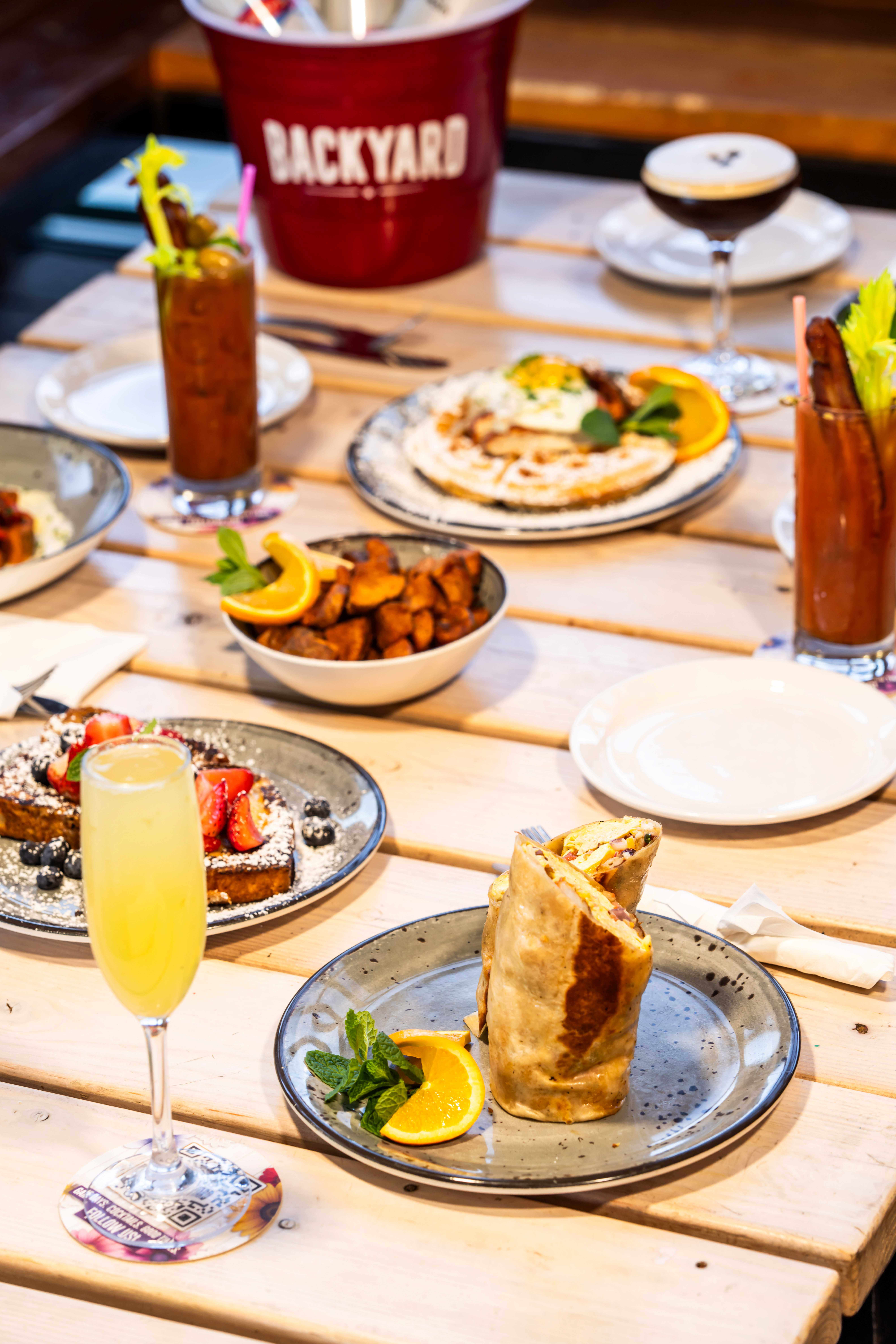 Brunch spread with breakfast dishes, cocktails, and a mimosa on a wooden patio table.
