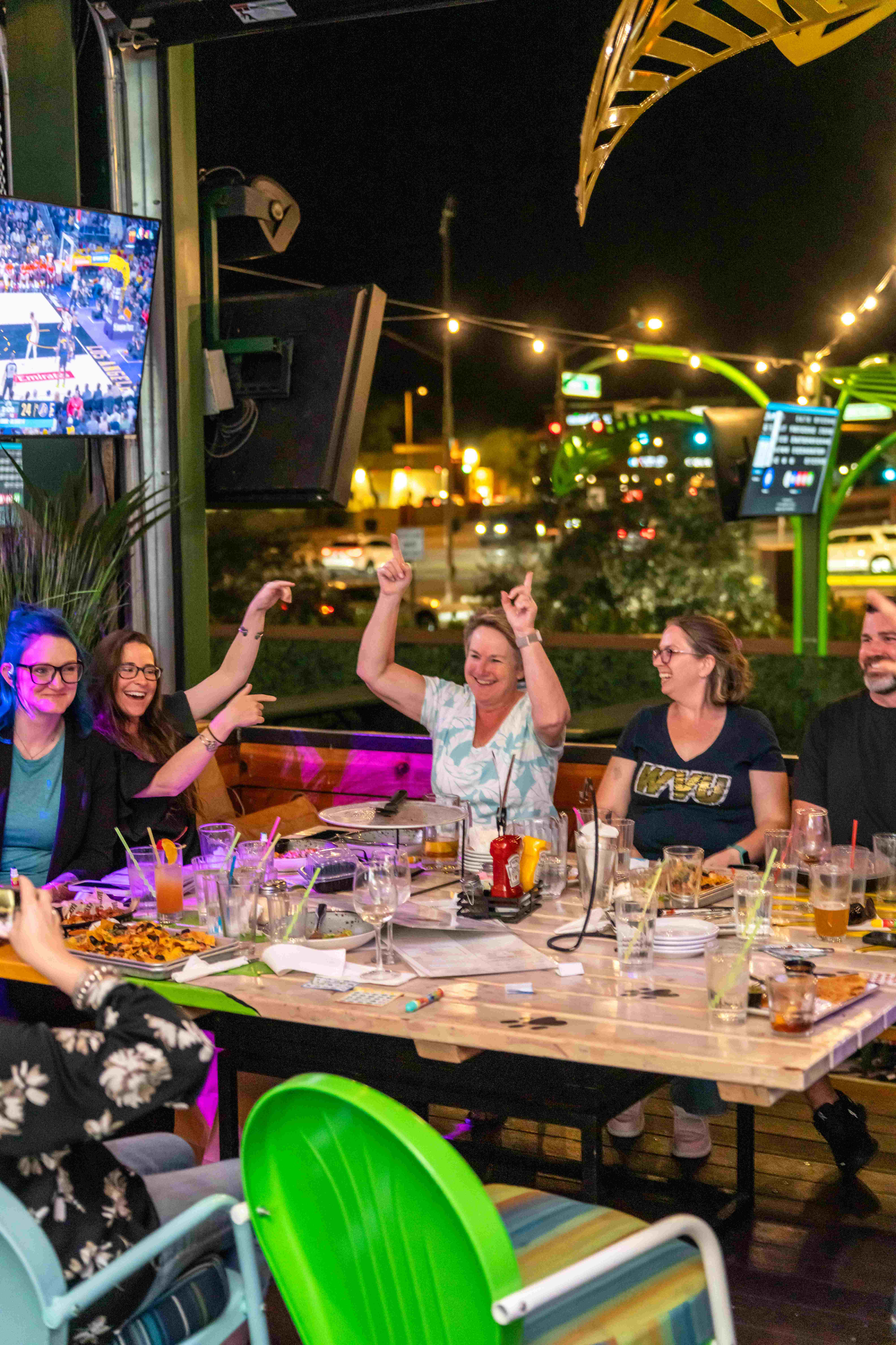 Group of people at an outdoor restaurant table at night pointing upward while playing bingo.