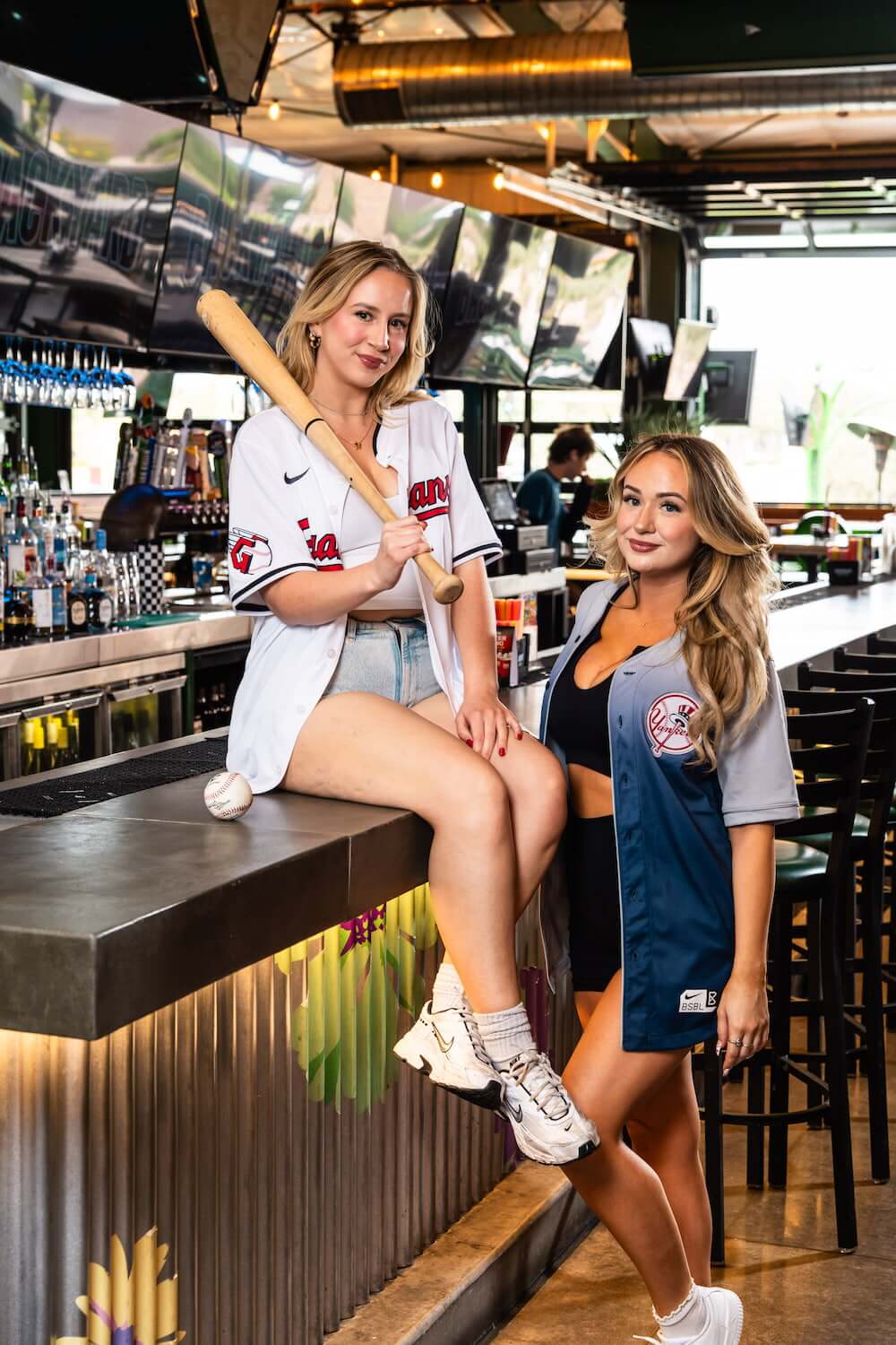 Two women in baseball jerseys posing in a bar, one holding a baseball bat and sitting on the counter