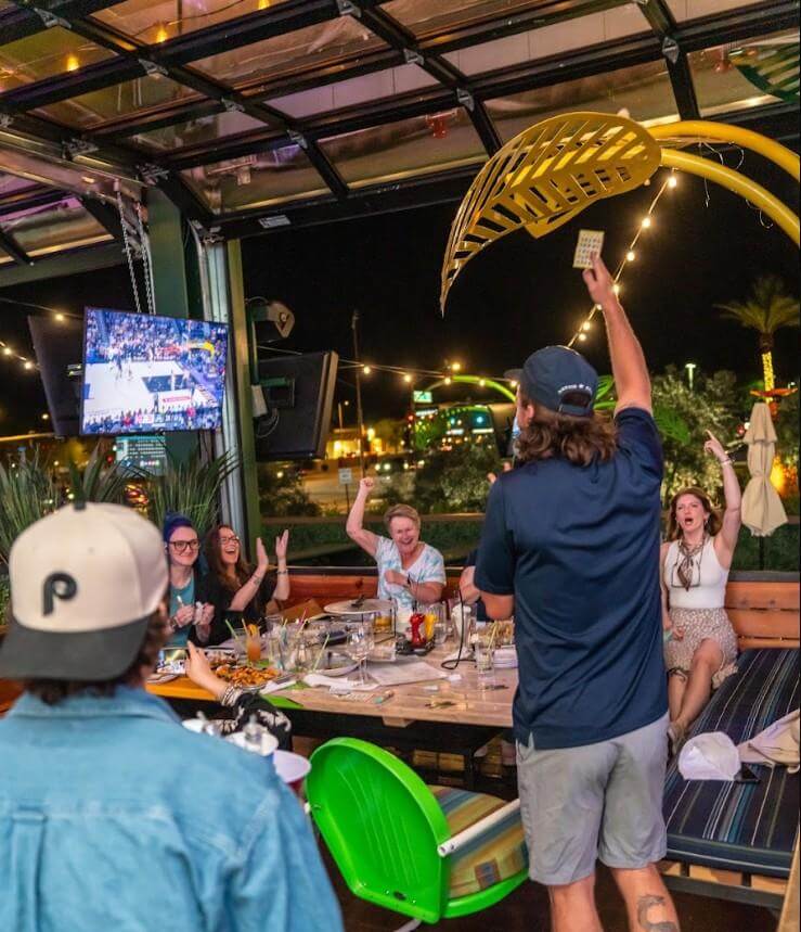 Guests cheer during a lively comedy bingo event at an outdoor sports bar, with a host calling numbers, a bingo card held in the air, and televisions showing a live basketball game in the background.