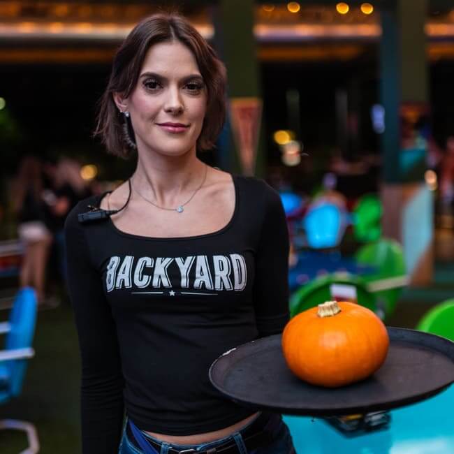 Backyard staff member holding a tray with a small orange pumpkin in a colorful outdoor dining area.