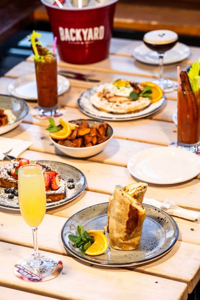 Brunch spread with breakfast dishes, cocktails, and a mimosa on a wooden patio table.