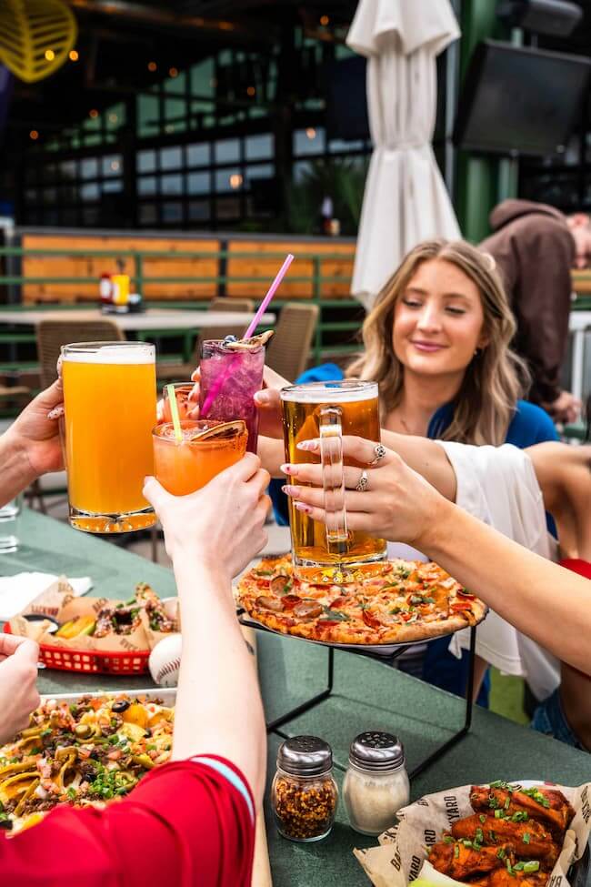 Group of friends clinking beers and cocktails over pizza and appetizers at an outdoor patio table.
